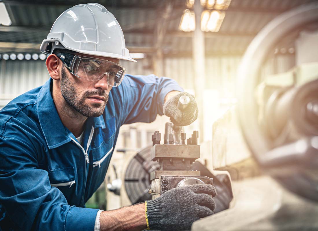 Manufacturer Insurance - Manufacturing Engineer Working in a Warehouse in a Blue Jumpsuit with a White Hard Hat and Operating a Lathe Machine
